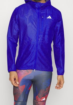 Mand iført en blå lynlåstrøje med hætte, hvidt logo og leggings med flerfarvet abstrakt mønster.