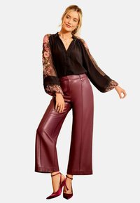 Blusa nera con maniche trasparenti a fiori, abbinata a pantaloni in ecopelle bordeaux a vita alta e tacchi viola. Design elegante ma moderno.