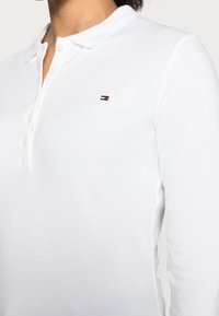 Hvid langærmet polo-shirt lavet af struktureret stof, med en knaplukket krave og et lille broderet logo i rød og blå på brystet.