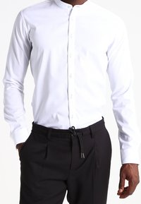 Vit långärmad skjorta med ståkrage, med knäppning framtill. Tillsammans med svarta byxor som har dragsko-detaljer.