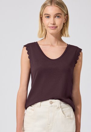 Donna bionda che indossa una canottiera scura con bordatura in pizzo e jeans a vita alta color avorio, in piedi davanti a uno sfondo chiaro e uniforme.