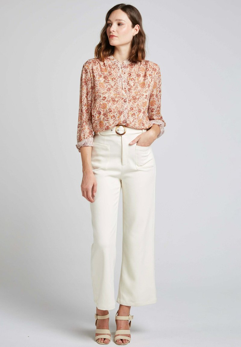 Blouse florale dans des tons chauds, col boutonné et manches retroussées, associée à un pantalon taille haute blanc avec une boucle de ceinture en or.