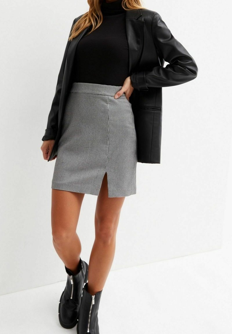 Femme portant un col roulé noir, un blazer en cuir noir, une mini-jupe grise à carreaux avec une fente à l'avant, et des bottines noires avec des fermetures éclair.