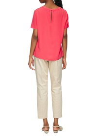 Kurzärmelige, leuchtend korallenfarbene Bluse mit Schlüsselloch-Rückendetail, kombiniert mit cremefarbenen, geraden Hosen und beige-farbenen Schuhen.