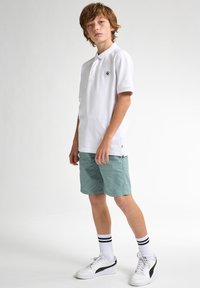 Chemise polo blanche avec un petit logo noir, accompagnée de shorts vert clair. Le modèle porte des baskets blanches avec des accents noirs et des chaussettes rayées.