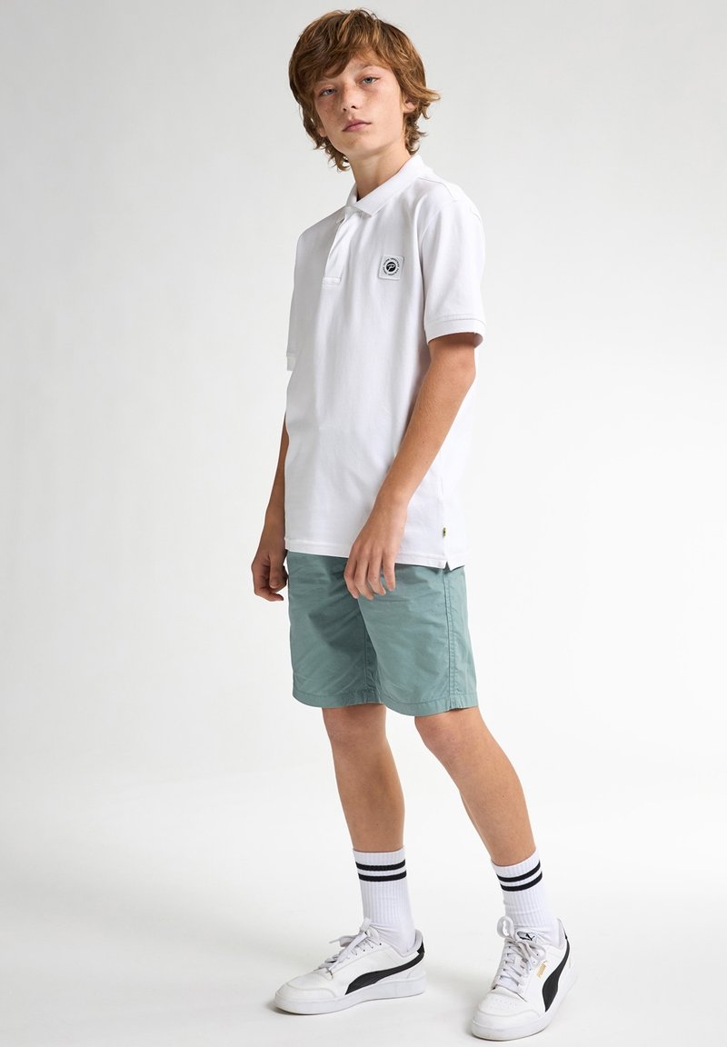 Chemise polo blanche avec un petit logo noir, accompagnée de shorts vert clair. Le modèle porte des baskets blanches avec des accents noirs et des chaussettes rayées.
