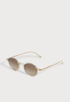 Lunettes de soleil rondes avec une monture claire translucide, des branches dorées et des verres bruns dégradés, sur un fond clair.