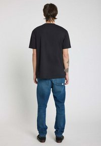 T-shirt noir à manches courtes avec un col rond, associé à un jean bleu en denim et des chaussures noires, vu de dos.