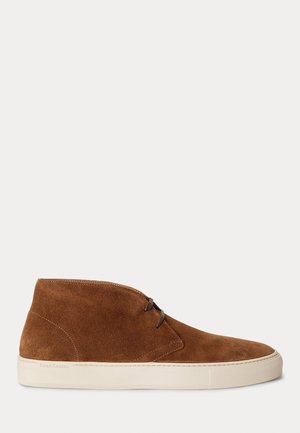 Brauner Wildleder-Mid-Top-Sneaker mit braunen Schnürsenkeln und weißer Gummisohle auf hellgrauem Hintergrund.