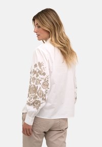 Bluză albă cu broderie florală bej pe mâneci, cu o croială lejeră și manșete cu nasturi. Textură netedă și material ușor.