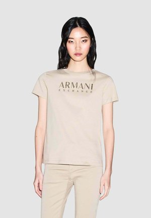 Donna che indossa una t-shirt beige Armani Exchange e pantaloni abbinati, in piedi davanti a uno sfondo semplice grigio chiaro.