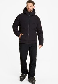 Schwarze Daunenjacke mit Reißverschluss vorne, Kapuze und zwei Seitentaschen, mit einem gesteppten Design und glatter Textur. Kombiniert mit schwarzen Hosen und Stiefeln.