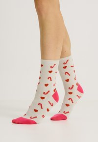 Calcetines gris claro con puntas y talones rosas, que presentan un patrón de corazones rojos y bastones de caramelo. Están hechos de un material suave y elástico.