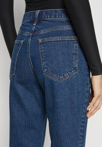 Jeans in denim blu scuro con vita alta, cinque tasche, cuciture visibili e leggermente sbiaditi. Il tessuto ha una consistenza liscia.