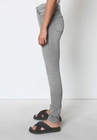 Jeans gris ajustados de cintura alta, con cinco bolsillos y detalles de costura, combinados con sandalias negras de tiras cruzadas sobre un fondo claro.