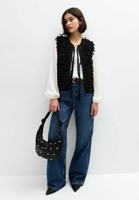 Zwarte textuurvest gelaagd over een witte blouse, gecombineerd met hoge taille blauwe denim flared jeans en een zwarte handtas met gouden accenten.
