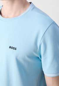 Chemise bleu clair à manches courtes avec des rayures blanches sur le col et "BOSS" imprimé en noir sur le côté gauche de la poitrine.