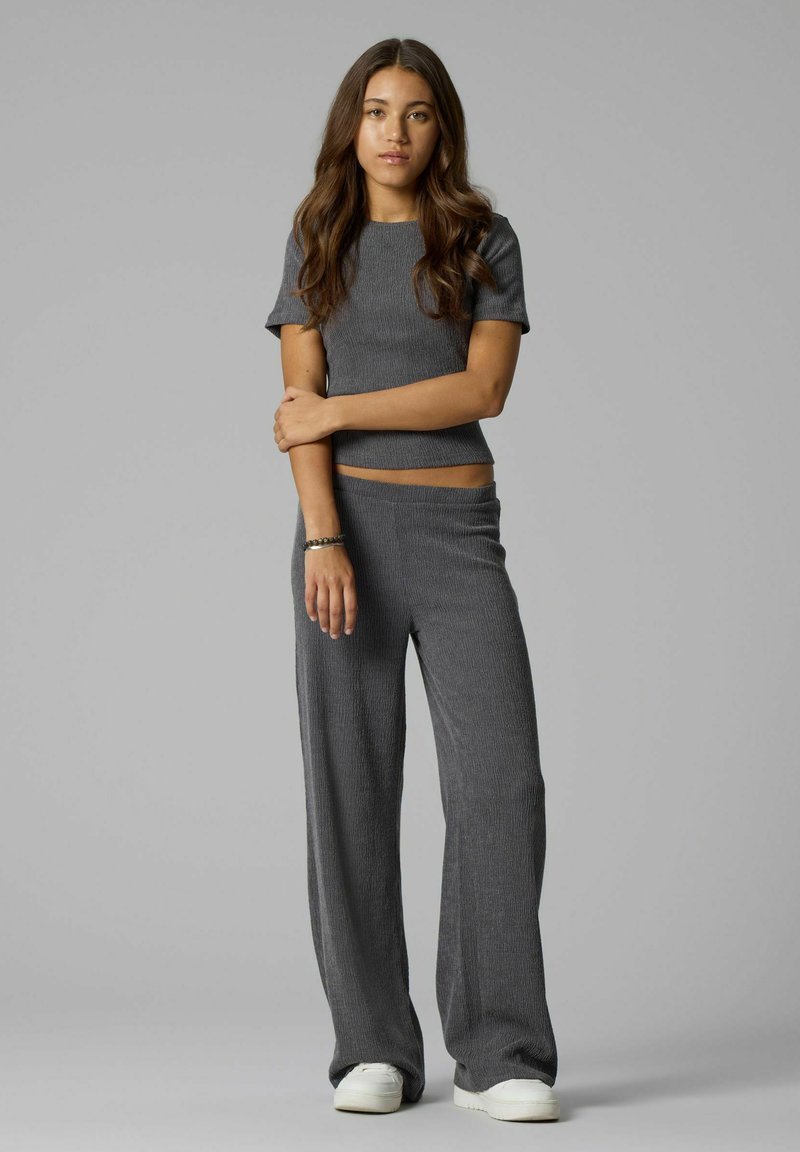 Jeune femme aux longs cheveux bruns portant un haut gris à manches courtes et un pantalon large assorti avec des baskets blanches sur fond gris.