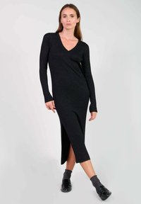 Robe en tricot côtelé noire avec un col en V, des manches longues et une fente latérale. Assortie avec des chaussettes grises et des chaussures noires, sur un fond neutre.