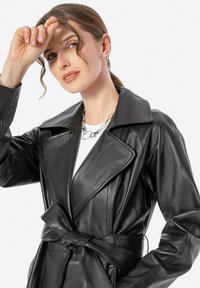 Cappotto trench in pelle nera con colletto ampio, vita cinturata e cuciture dettagliate. Il modello indossa una collana di catena argentata.