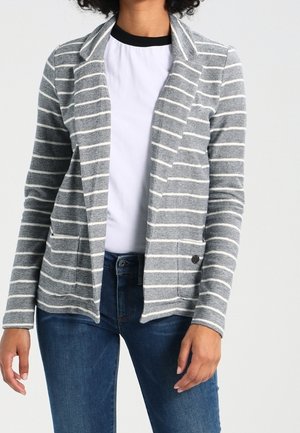 Nainen pukeutunut harmaavalkoraidalliseen avoimeen bleiseriin valkoisen paidan mustalla kauluksella ja sinisten farkkujen kanssa yksiväristä taustaa vasten.