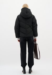 Juodas pūkų paltas su gobtuvu, quilted dizainas ir elastingi rankogaliai. Suderintas su tamsiomis džinsomis ir rudų spalvos rankine. Fone paprasta aplinka.