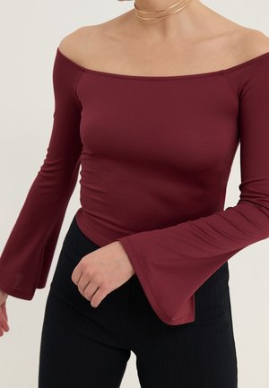 Kobieta nosząca bordowy top z opuszczonymi ramionami i długimi rękawami z rozszerzanymi mankietami oraz czarne prążkowane spodnie, z złotym naszyjnikiem choker.