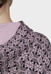 Mikina s kapucí se geometrickým černobílým a růžovým vzorem. Tkanina má hladkou strukturu, s viditelnou světle fialovou podšívkou.