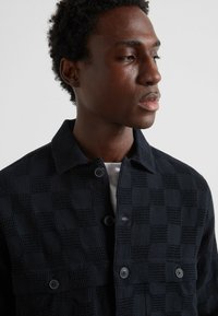 Giacca a camicia a scacchi navy realizzata in tessuto a maglia testurizzato con chiusura a bottone, dotata di due tasche frontali e un colletto classico.