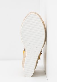 Semelle de sandale compensée avec dessous blanc texturé, côtés en tissu beige tissé et bride à boucle jaune, positionnée verticalement contre des murs d'angle.