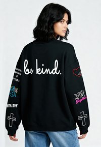 Čierna oversize mikina s bielym nápisom "buď láskavý." na chrbte, farebnými grafickými dizajnmi na rukávoch a rebrovanými manžetami.