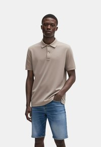 Polo beige con collo a polo, due bottoni, maniche corte e un logo discreto, abbinato a pantaloni corti in denim azzurro chiaro.