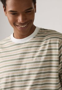 Rutet T-skjorte med horisontale linjer i grønn og beige, med hvit rundhals. Diskret logo brodert på brystet.