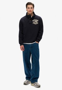Granatowy sweter z wysokim kołnierzem, z zamknięciem na guziki z przodu oraz białym logo "SUPERDRY JPN ATHLETIC". Noszony z niebieskimi dżinsami.