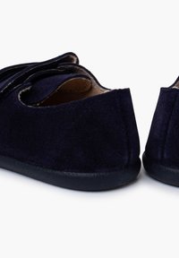 Zapatillas de color azul marino hechas de un suave tejido con una suela plana de goma negra. Presentan una punta redondeada y un diseño cosido a lo largo de la parte trasera.