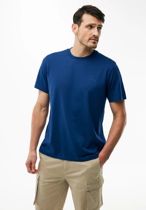 Uomo che indossa una maglietta blu tinta unita a maniche corte e pantaloncini cargo beige, in piedi con una mano in tasca, guardando di lato.