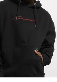 Sweat à capuche noir avec le texte "Rocawear" brodé en rouge sur la poitrine. Comprend des cordons de serrage, une poche avant, des poignets et un ourlet côtelés. Texture douce.