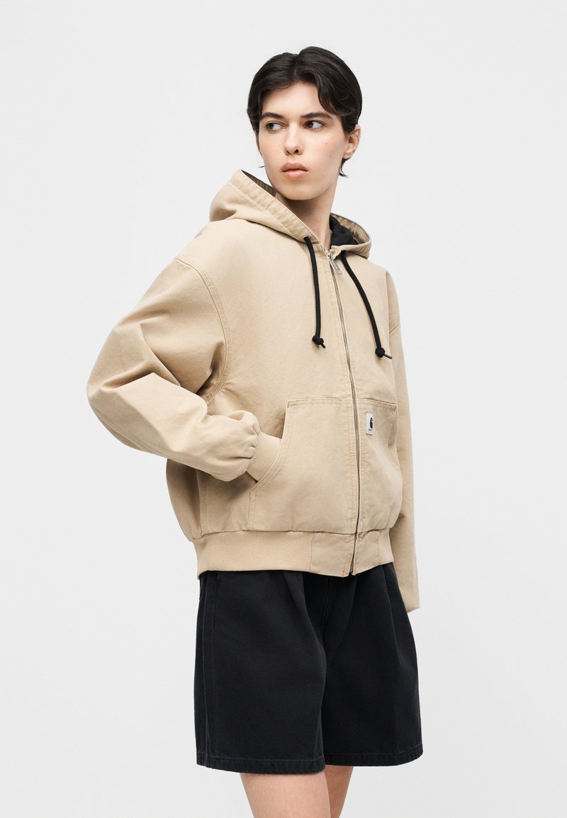 Jeune personne portant un sweat à capuche beige zippé avec des cordons noirs et un short noir jusqu'aux genoux, regardant sur le côté avec la main dans la poche.