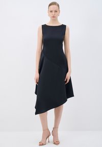 Robe noire sans manches avec un corsage ajusté, un ourlet asymétrique et une texture satinée lisse. Associée à des escarpins à brides nude.