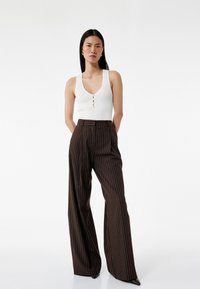 Witte geribbelde tanktop met een V-hals en drie knoopdetails, gecombineerd met bruine high-waisted broek met pinstripes. Soepele texturen en brede pijpen.