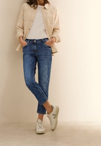 Giacca button-up beige chiaro, t-shirt bianca, jeans di denim blu e sneakers bianche con dettagli grigi, con una vestibilità rilassata e orli arrotolati.