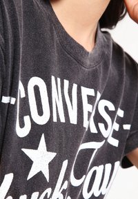 T-shirt noir en coton avec col rond, présentant un grand texte blanc "CONVERSE" et un graphique d'étoile, à texture légèrement usée.