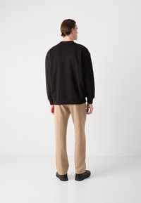 Crni oversized dukserica s rebrastim manžetama i rubom, uparen s bež hlačama. Model okrenut leđima, nosi crne cipele. Minimalistička pozadina.