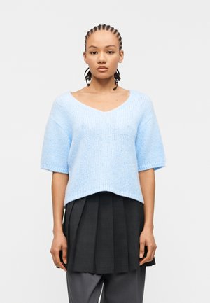 Žena s copánky, která má na sobě světle modrý krátký pletený svetr a vrstvenou černou plisovanou sukni přes šedé kalhoty na bílém pozadí.