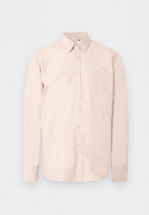 Chemise rose clair à manches longues avec boutons, col pointu, logo discret sur la poitrine et poignets boutonnés sur un fond blanc uni.