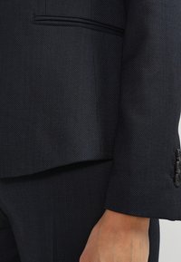 Svart texturerad blazer med slim fit, med en enkel bröstficka och tre-knappsfäste vid ärmarna. Slätt tyg med subtilt mönster.