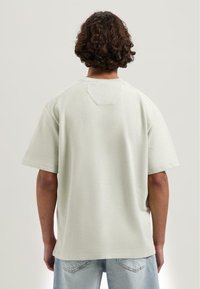 Cămașă de bumbac de culoare verde deschis, cu mâneci scurte, decolteu rotund și material texturat. Conține un detaliu mic sub formă de patch în apropierea decolteului.