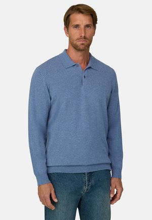 Homme aux cheveux châtain clair portant un pull polo bleu à manches longues et un jean bleu, debout devant un fond blanc uni.