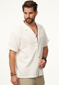 Weißes Kurzarmhemd mit strukturiertem Blumenmuster, mit eingekerbtem Kragen und braunen Knöpfen, kombiniert mit hellen, strukturierten Hosen und Accessoires.