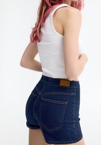Mujer con cabello rosa que lleva una blusa sin mangas blanca y pantalones cortos de mezclilla azul oscuro, vista de perfil sobre un fondo liso.